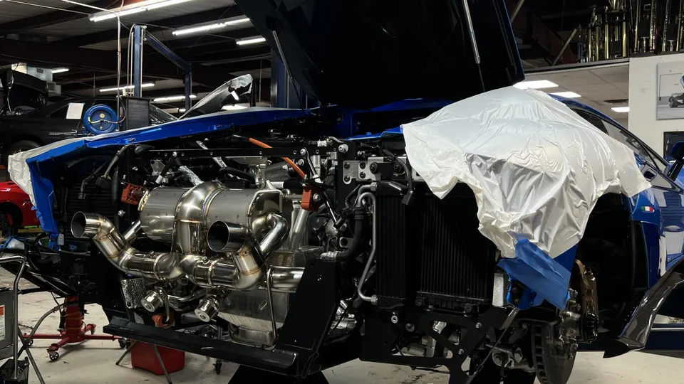 A blue Lamborghini supercar in a shop with its engine bay exposed, showcasing a custom stainless steel exhaust system.