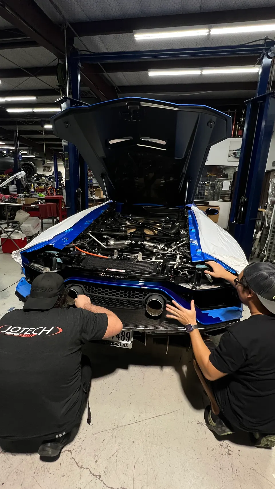 Two mechanics work on the rear exhaust and open engine bay of a blue Lamborghini Huracan in a Jotech Motorsports shop.