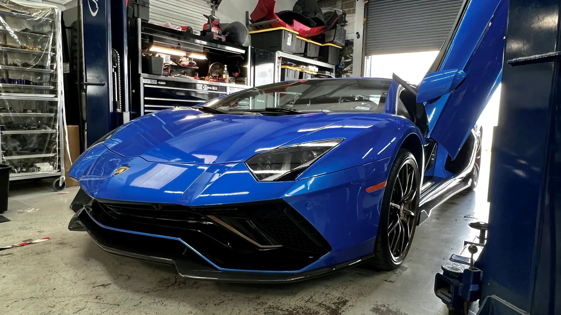 A blue Lamborghini Aventador with its scissor doors open, parked inside a performance tuning shop.