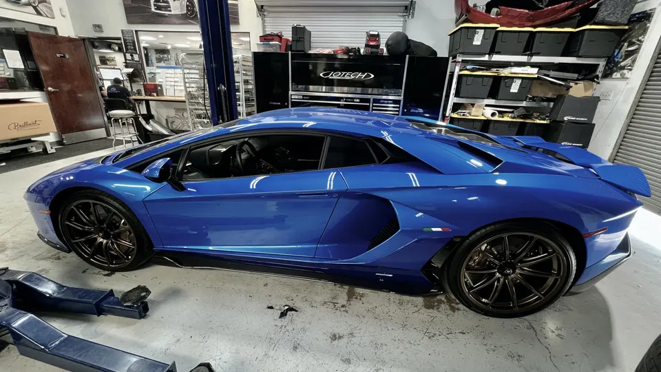 A blue Lamborghini Aventador supercar parked inside a performance tuning shop with tools and equipment in the background.