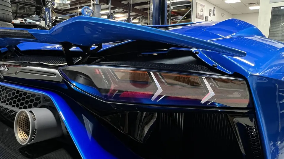 Blue Lamborghini rear with a large wing, distinctive taillight, and performance exhaust tip in a shop setting.
