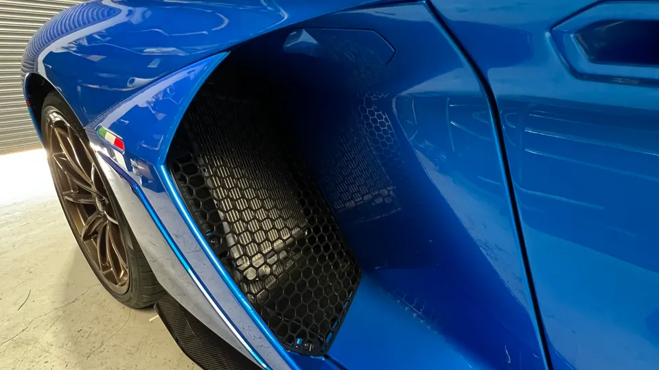 A close-up detail of a blue Lamborghini's side air intake, fender, and bronze wheel with an Italian flag badge.
