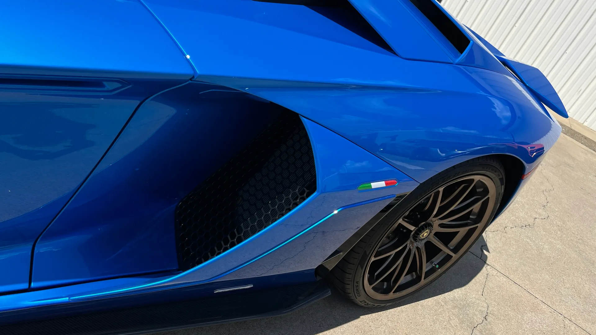 A close-up of a blue Lamborghini Aventador's rear quarter panel, showing the side air intake, bronze wheel, and Italian flag badge.