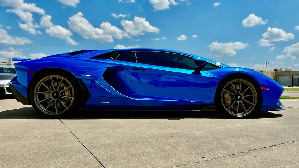 A blue Lamborghini Aventador parked outdoors under a clear sky, viewed from the side.