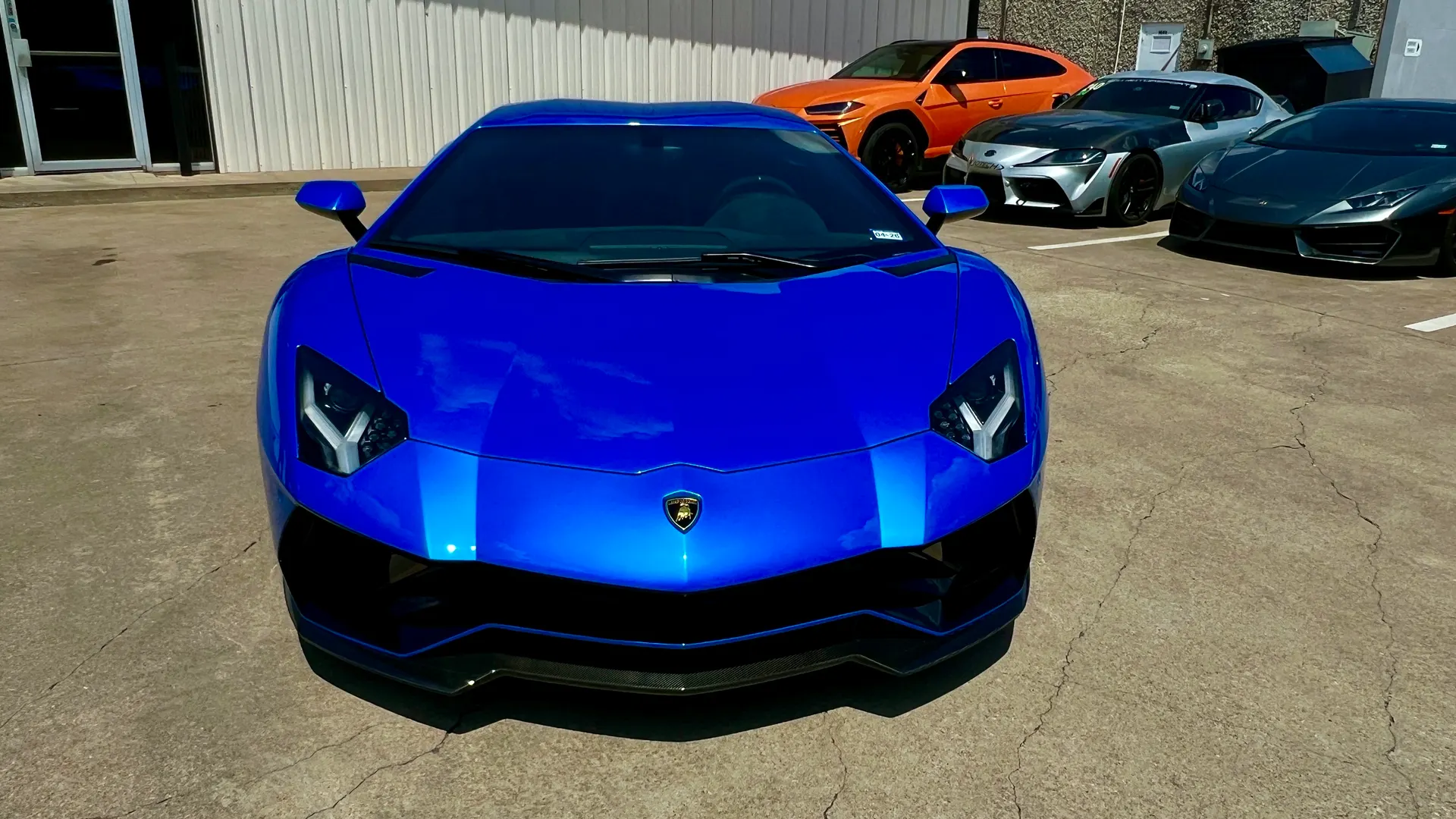 A vibrant blue Lamborghini Aventador parked prominently in a lot with other luxury cars, including an orange Lamborghini Urus and a silver Toyota Supra.