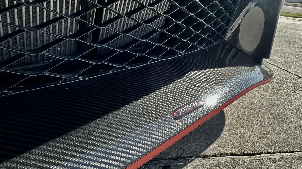 Close-up of a Jotech carbon fiber front splitter on a performance car, showcasing the weave and logo.