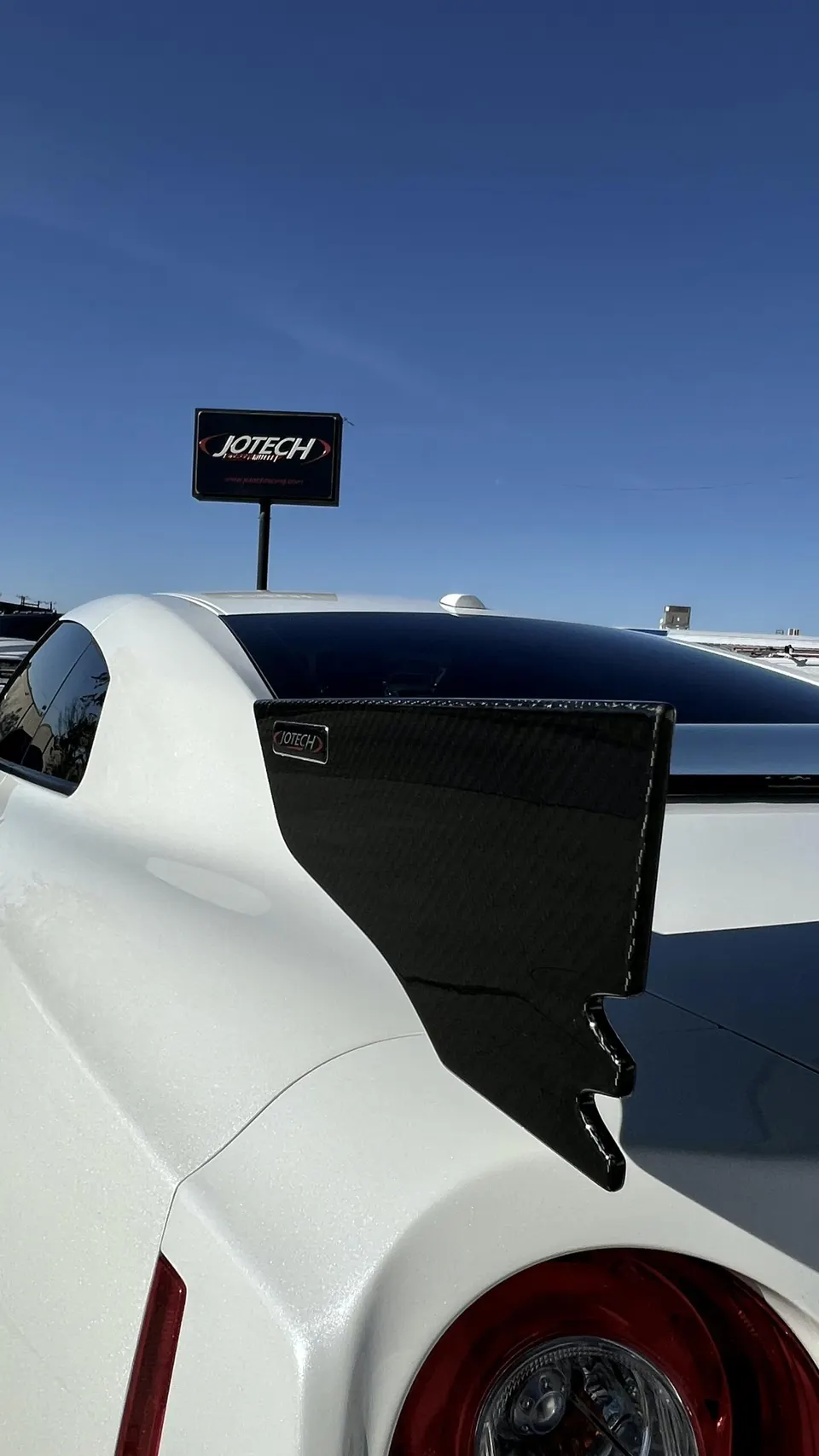 Rear view of a white Nissan GT-R featuring a carbon fiber wing, with a Jotech sign visible in the background.