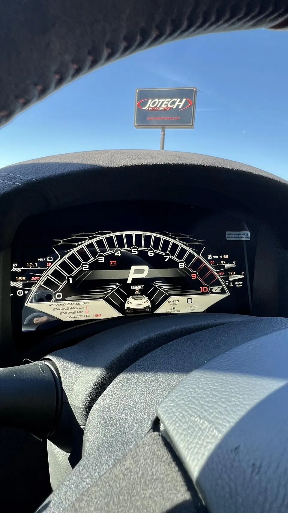 A view from inside a car showing a digital dashboard with various gauges and a Jotech Motorsports sign visible through the windshield.