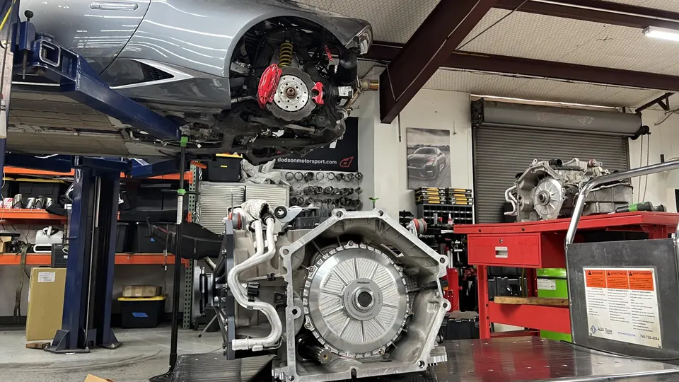 A silver car on a lift in a performance shop with a transmission on a table in the foreground, showing work in progress.