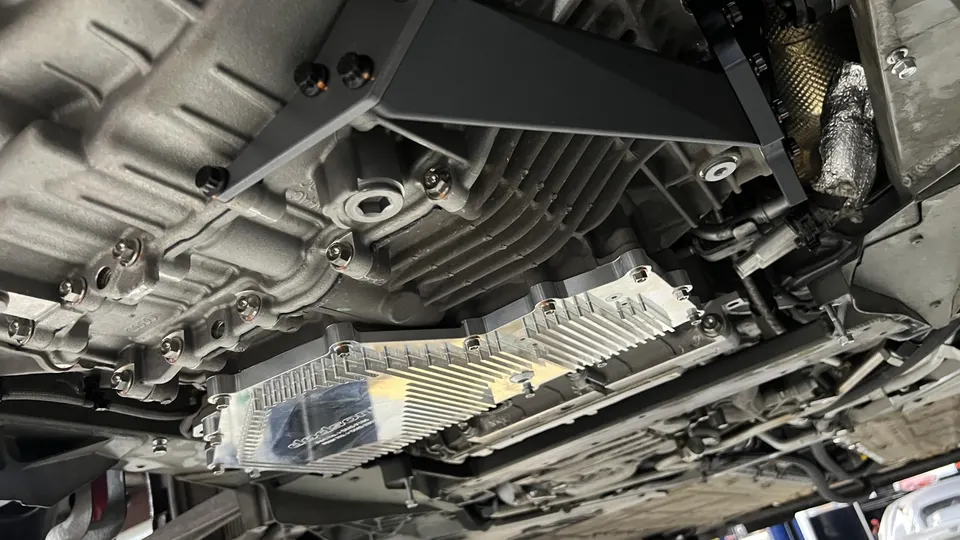 Underside view of a performance car's drivetrain, showing the transmission and an aftermarket cooling pan in a shop.