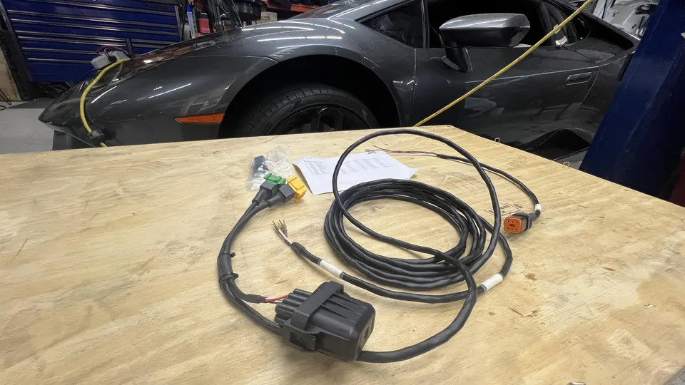 Wiring harness and connectors on a wooden table in front of a grey Lamborghini Huracan in a performance tuning shop.