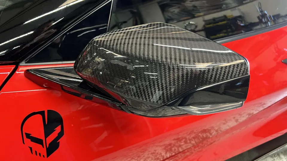 Close-up of a carbon fiber side mirror on a red Corvette, showcasing the weave pattern and glossy finish.
