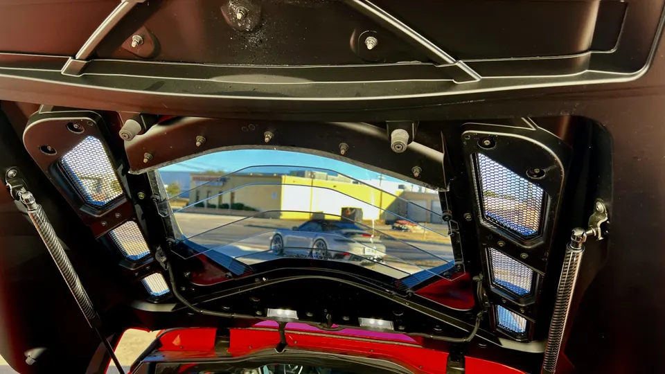 Underside of a car's hood with performance vents and carbon fiber struts, looking out at a parked Porsche.