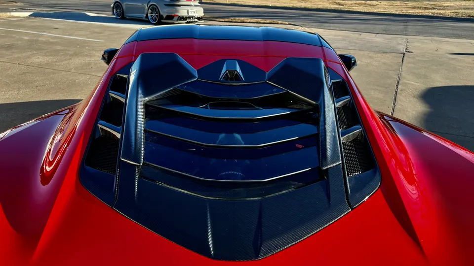High-angle view of a red Corvette C8's rear engine cover with carbon fiber louvers and vents, parked outdoors.