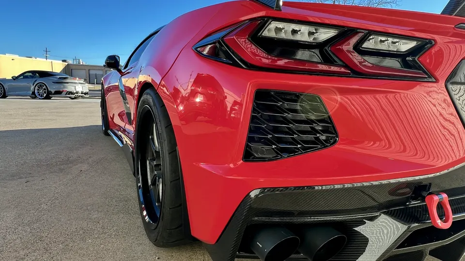 A low-angle rear view of a red C8 Corvette with a carbon fiber diffuser and exhaust tips, parked outdoors with a grey Porsche 911 in the background.