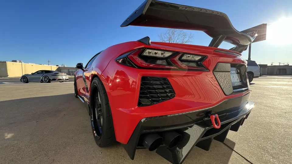 Red C8 Corvette with a large carbon fiber wing and aggressive rear diffuser, parked outdoors with a grey Porsche in the background.