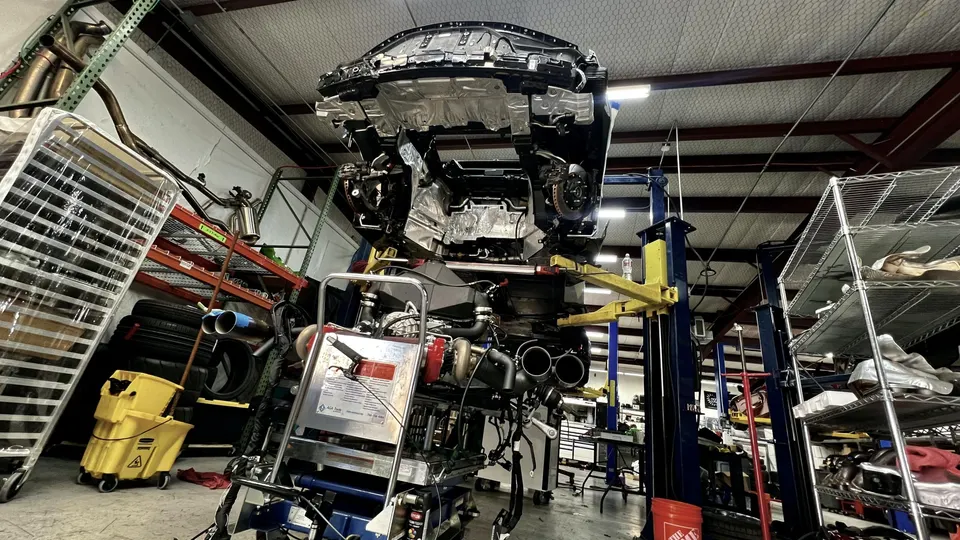 A car undergoing a performance build in a shop, with the chassis on a lift and the engine/drivetrain assembly on a cart.