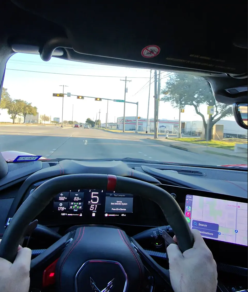 Driver's view from inside a red Corvette C8, showing the steering wheel, digital dashboard, and road ahead.