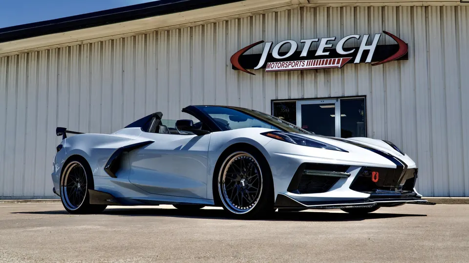 A white Corvette C8 convertible with aftermarket wheels, a large rear wing, and a front splitter, parked outside Jotech Motorsports.