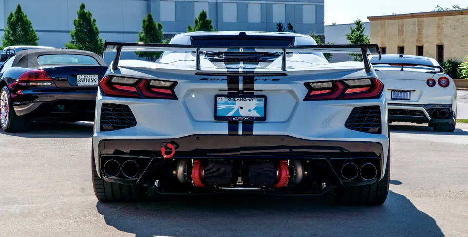 Rear view of a white Corvette C8 with exposed twin turbos and a large wing, parked next to a black Dodge Viper and a white Nissan GT-R.