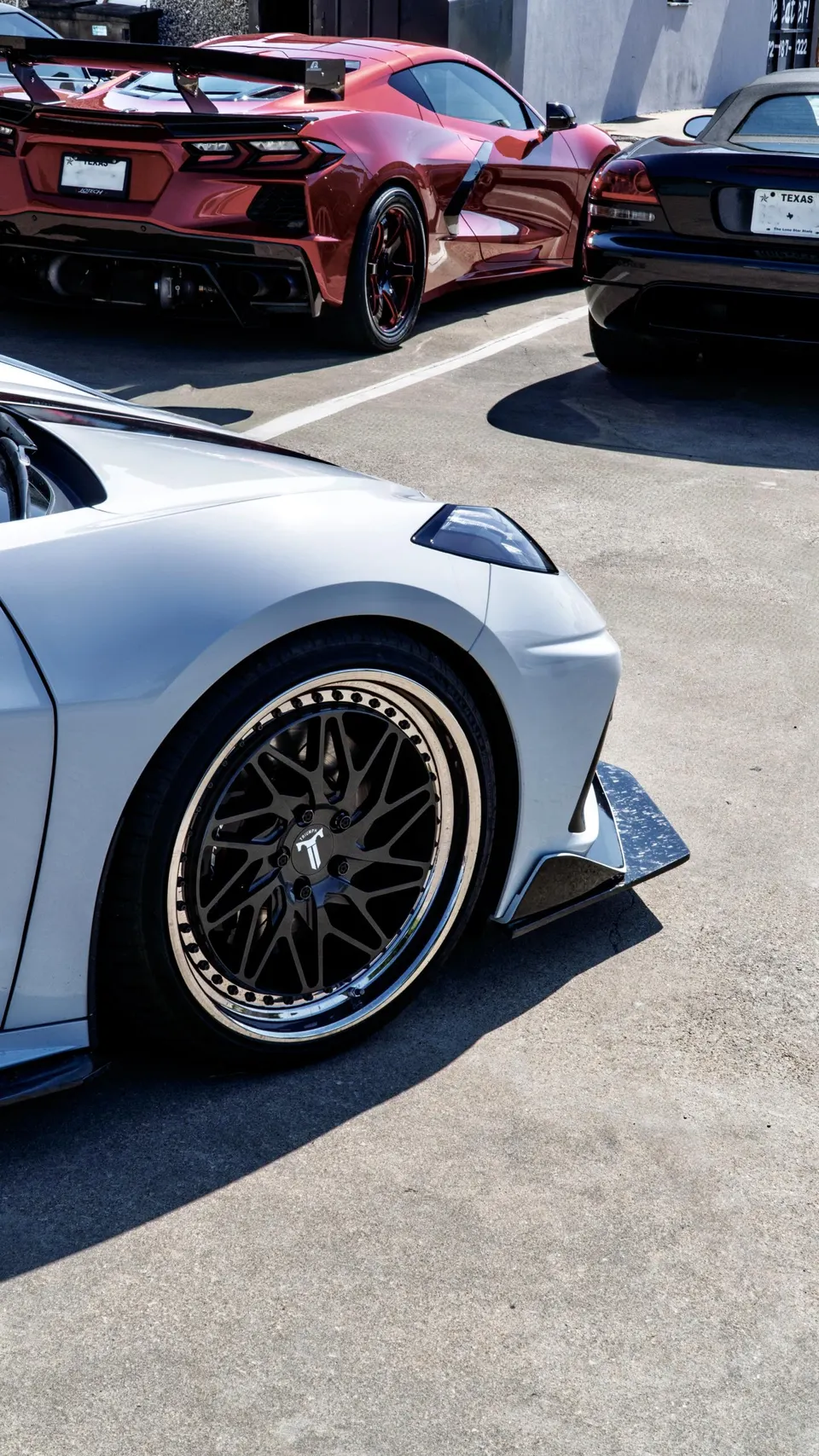Close-up of a white Corvette C8 with custom black forged wheels and a carbon fiber front splitter, parked outdoors.