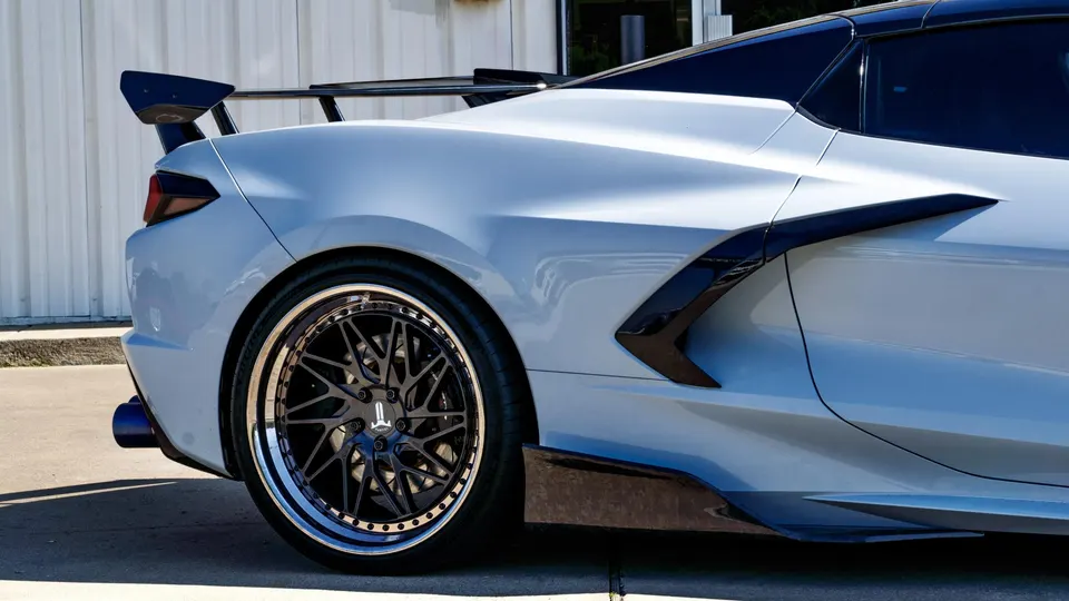 Rear quarter view of a grey Corvette C8 with a large aftermarket rear wing, custom black wheels, and side skirts.