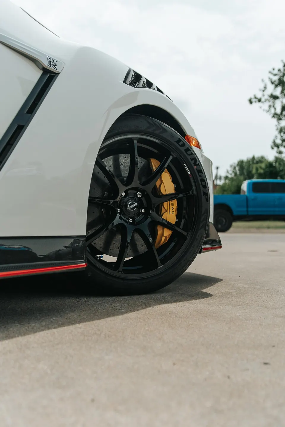 Close-up of a white Nissan GT-R's front wheel, featuring black multi-spoke wheels, yellow brake calipers, and Dunlop tires.