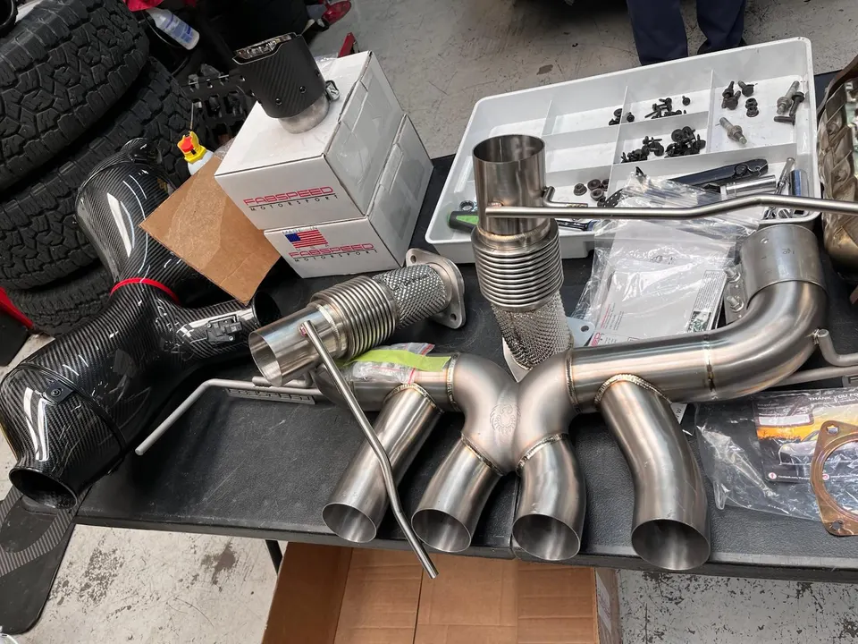 Performance car parts laid out on a table, including a Fabspeed carbon fiber intake and stainless steel exhaust components.
