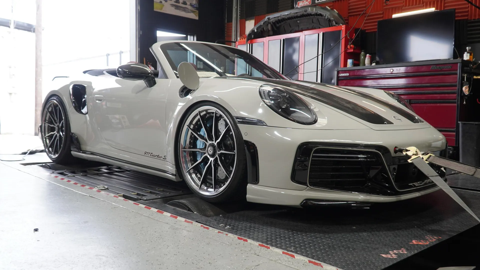 A white Porsche 911 Turbo S convertible strapped to a dynamometer in a performance tuning shop.