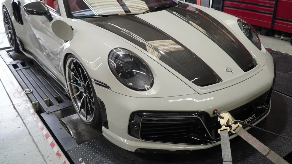 A white Porsche 911 Turbo S with carbon fiber stripes strapped down on a dynamometer for performance testing.