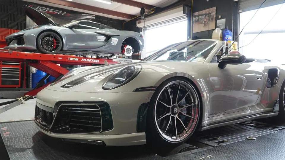 A grey Porsche 911 Turbo S on a dyno with a grey Corvette C8 on a lift in a performance tuning shop.