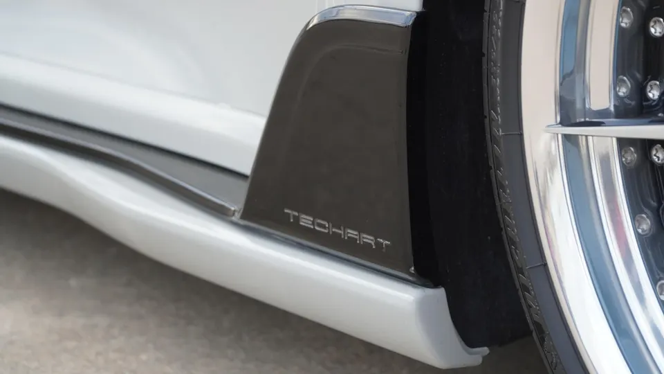 A close-up of a white car's side skirt with "TECHART" branding and a chrome multi-spoke wheel.