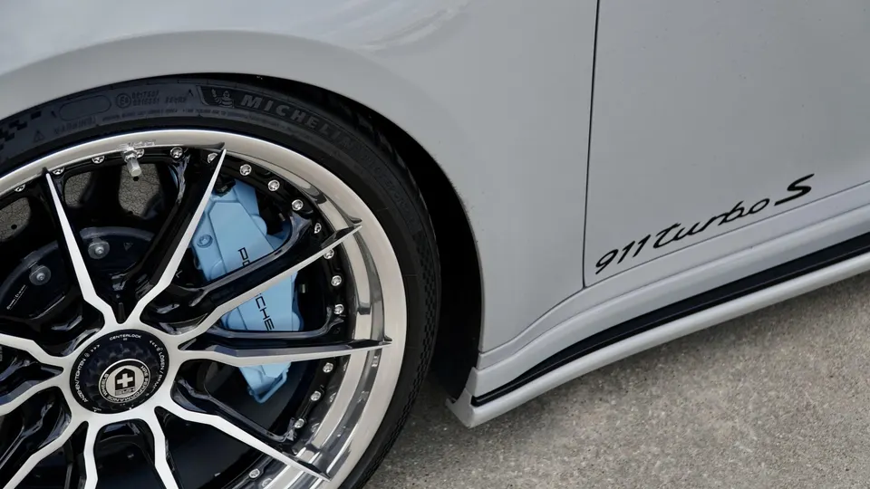 A close-up of a silver Porsche 911 Turbo S wheel with black and chrome spokes and light blue brake calipers.