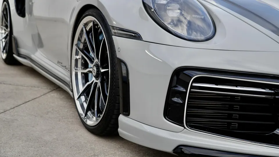 Close-up of a light grey Porsche 911 Turbo S showing custom multi-spoke wheels, front fender, and headlight.