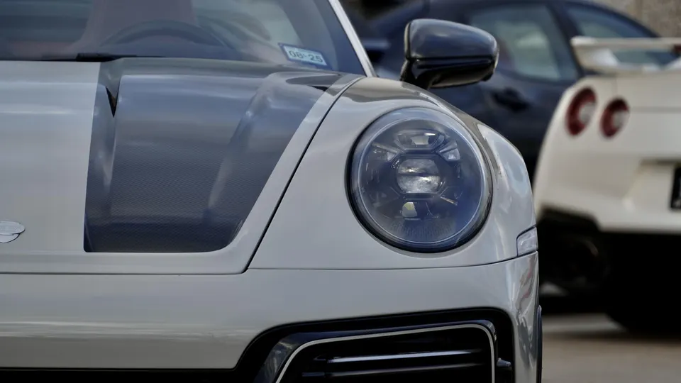 Front view of a white Porsche 911 with a carbon fiber hood stripe, parked with a white Nissan GT-R in the background.