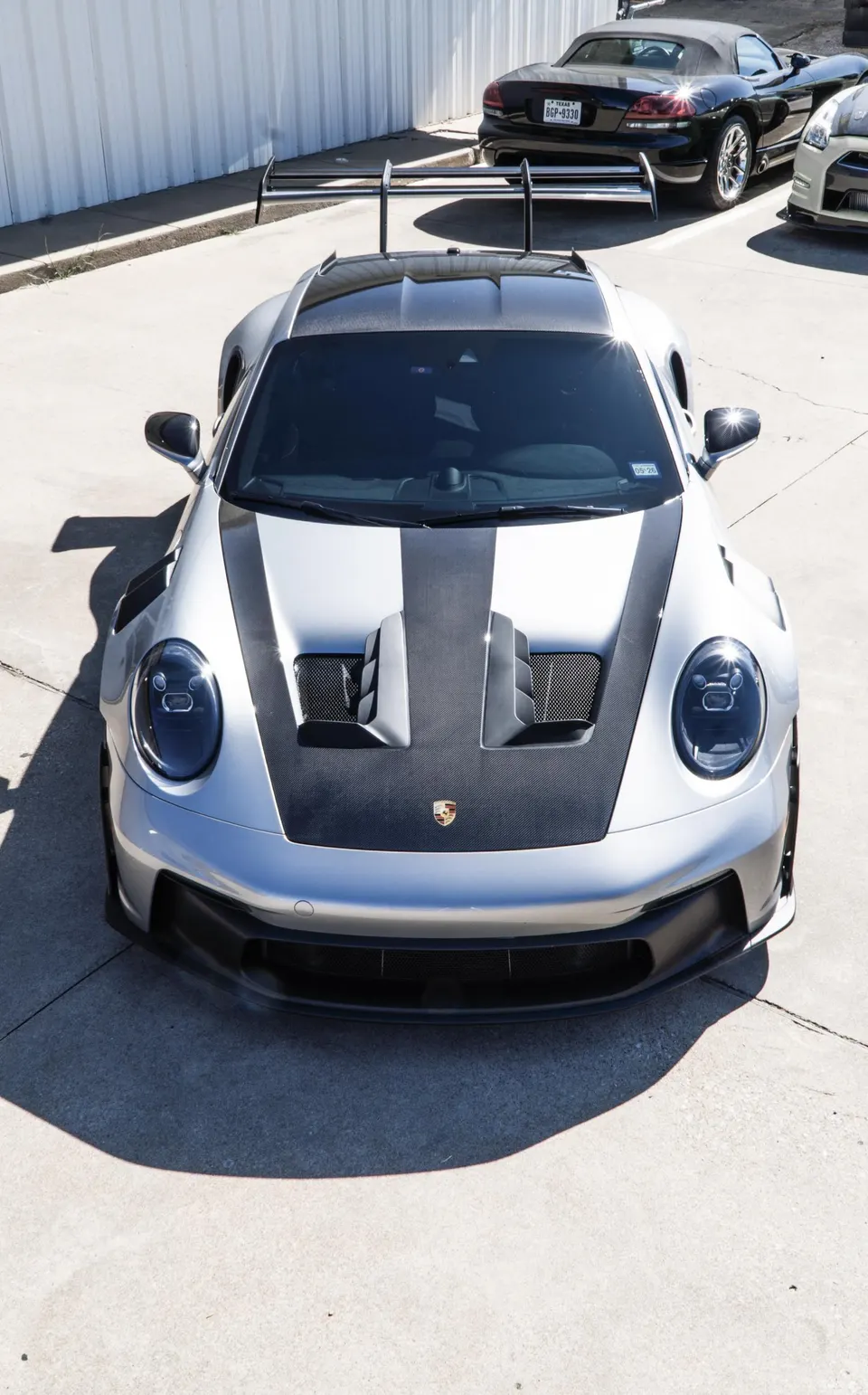 Silver Porsche 911 GT3 RS with a carbon fiber hood and large rear wing parked outdoors on a sunny day.