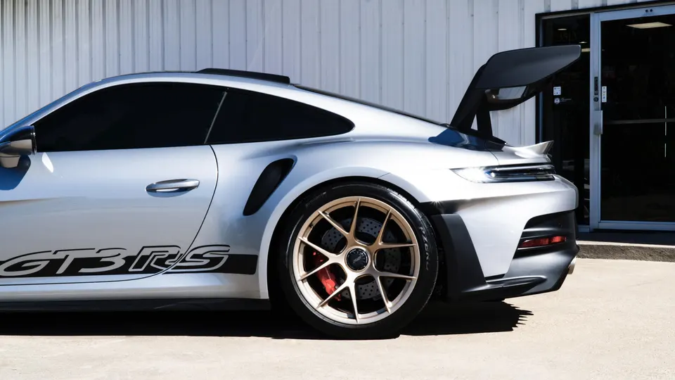 Side rear view of a silver Porsche 911 GT3 RS with gold aftermarket wheels and a large black rear wing.