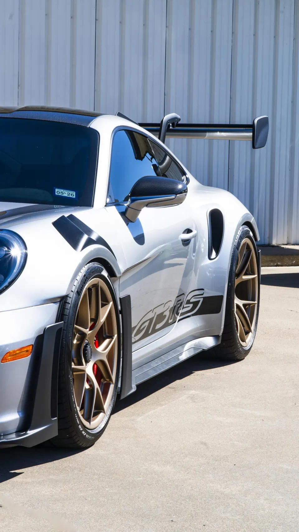 Silver Porsche 911 GT3 RS with gold wheels and large rear wing parked outdoors against a metal wall.