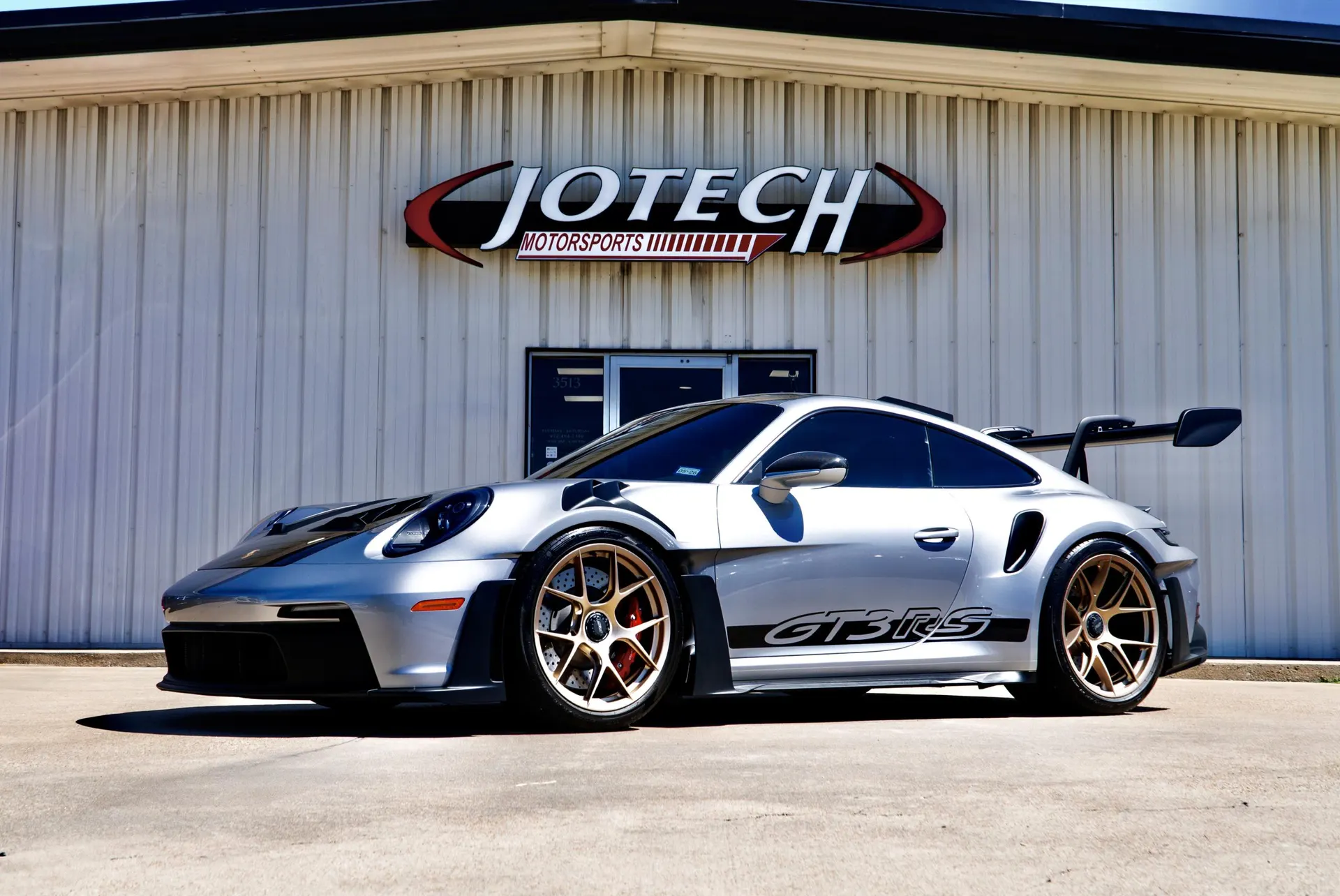 Silver Porsche 911 GT3 RS with gold aftermarket wheels and large rear wing parked in front of Jotech Motorsports shop.