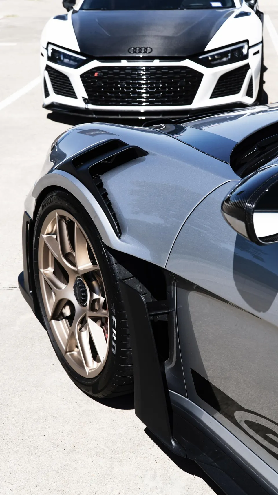 Close-up of a grey sports car with gold wheels and carbon fiber accents, with a white Audi R8 in the background.