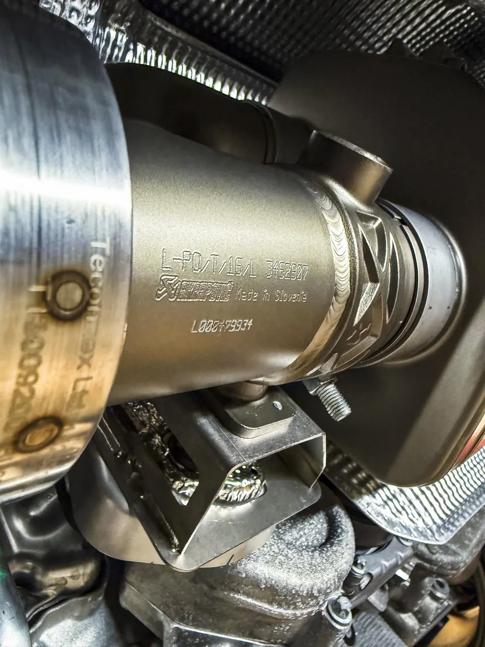 Close-up of an Akrapovic titanium exhaust downpipe with visible welds and part numbers installed on a performance car.