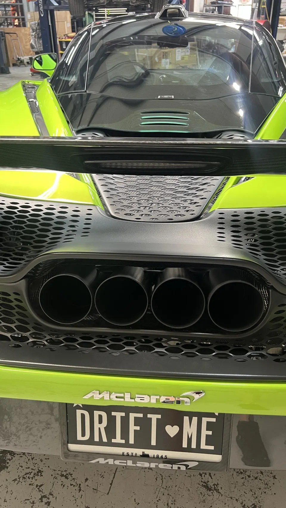 Rear view of a bright green McLaren with quad exhaust tips, a black rear diffuser, and a custom 'DRIFT ME' license plate in a shop.