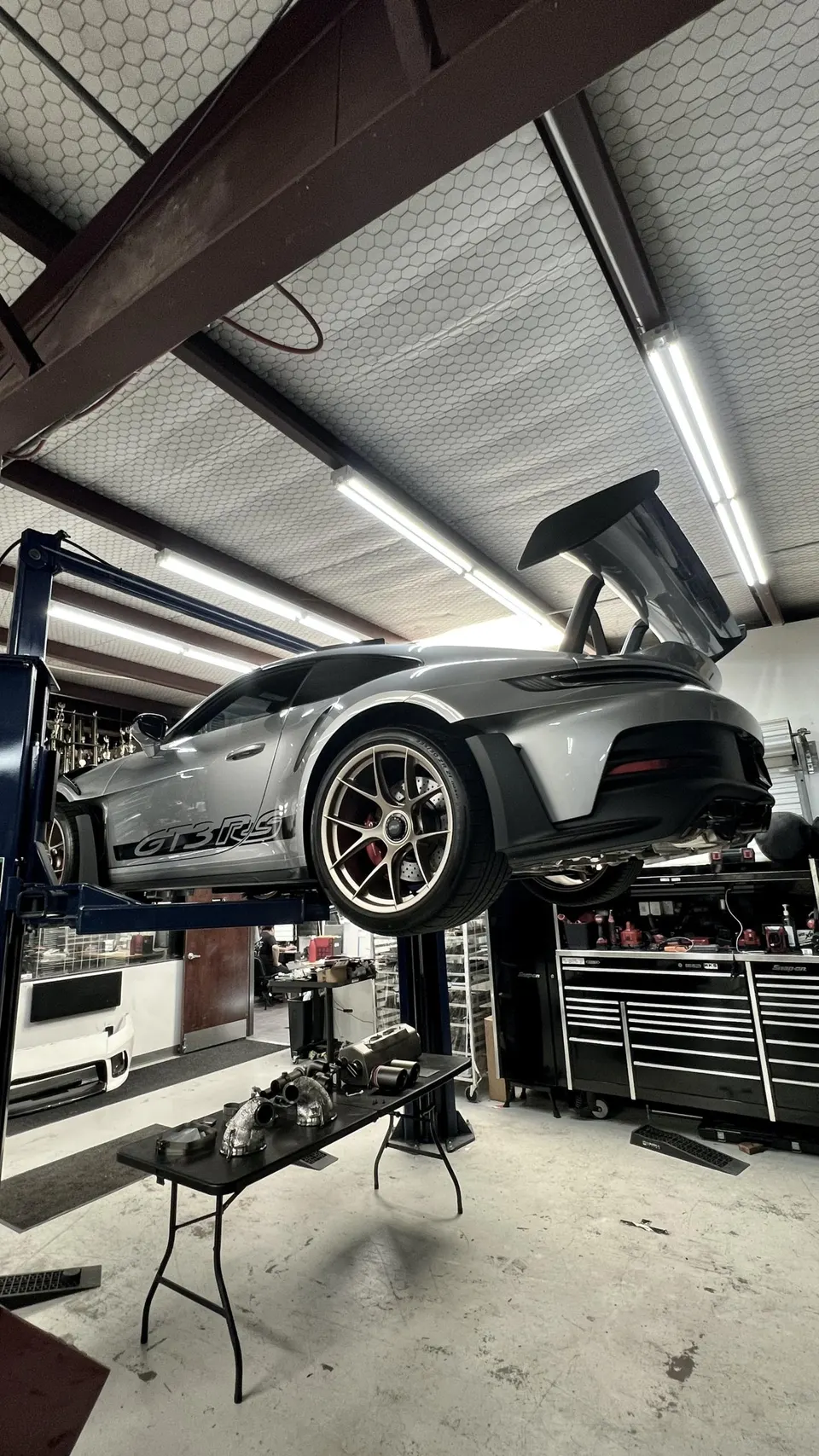 A silver Porsche GT3 RS on a lift in a performance tuning shop with turbochargers and other parts on a table below.