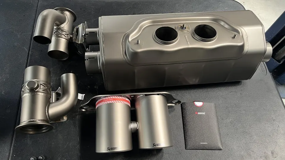 Akrapovic titanium exhaust system components including muffler, connecting pipes, and exhaust tips laid out on a table.
