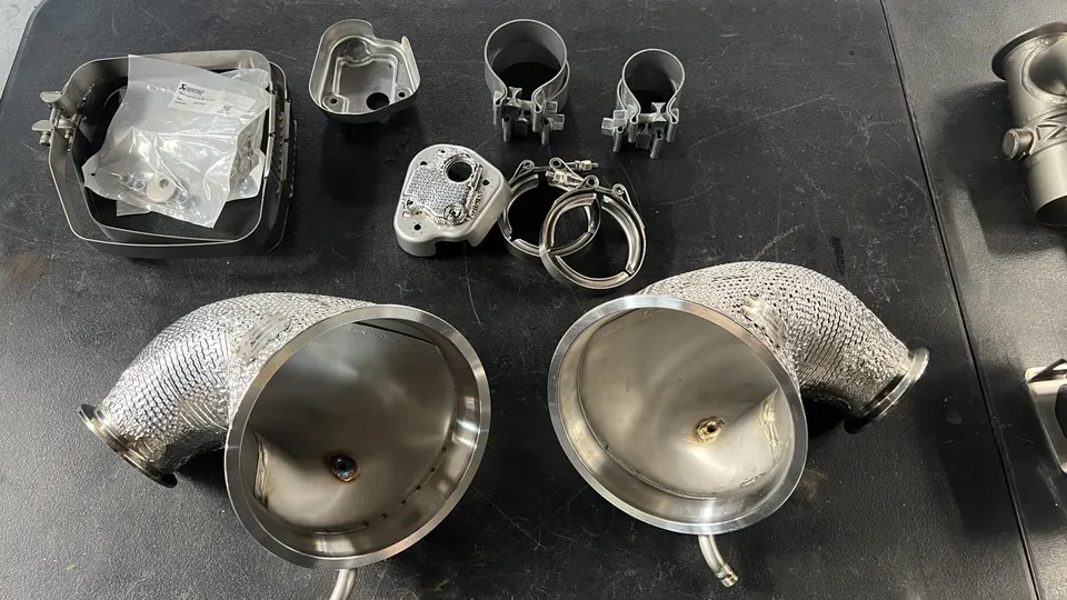 Akrapovic exhaust system components including heat-shielded downpipes and clamps laid out on a workbench.