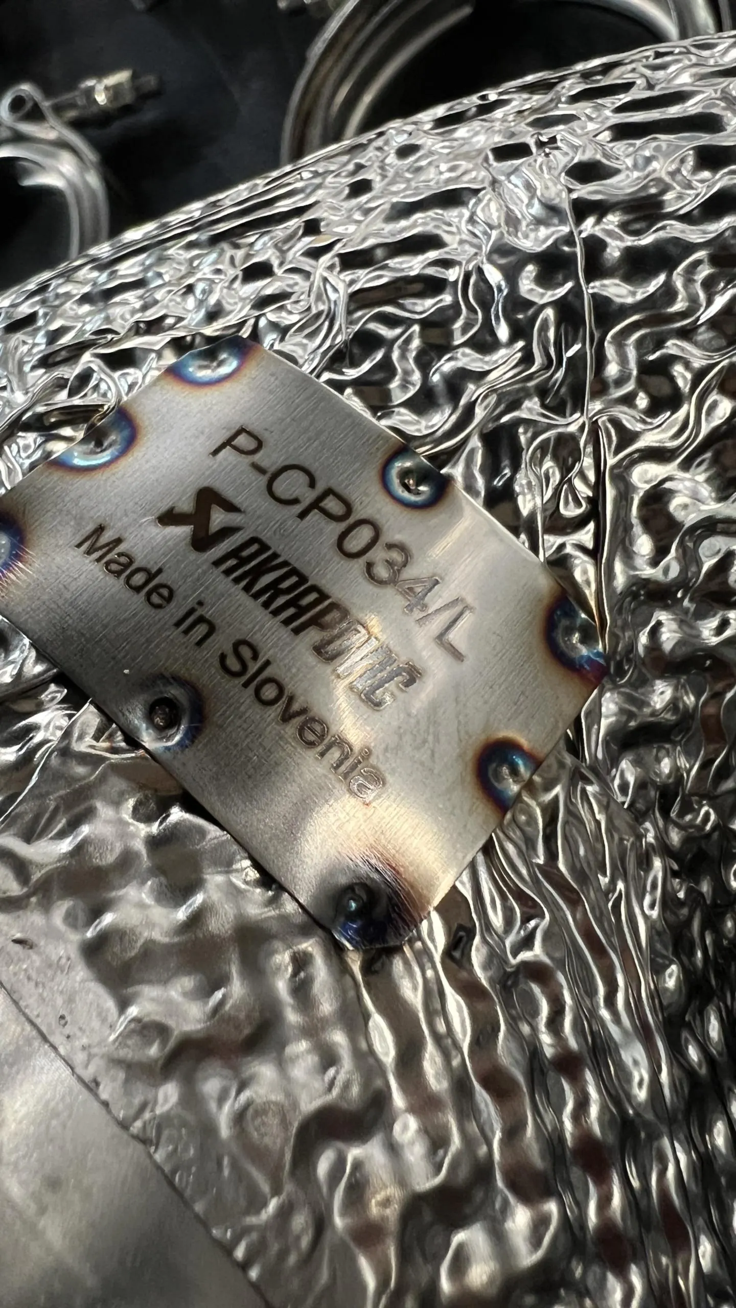 Close-up of an Akrapovic exhaust component with part number P-CP034/L and "Made in Slovenia" on a metal tag.