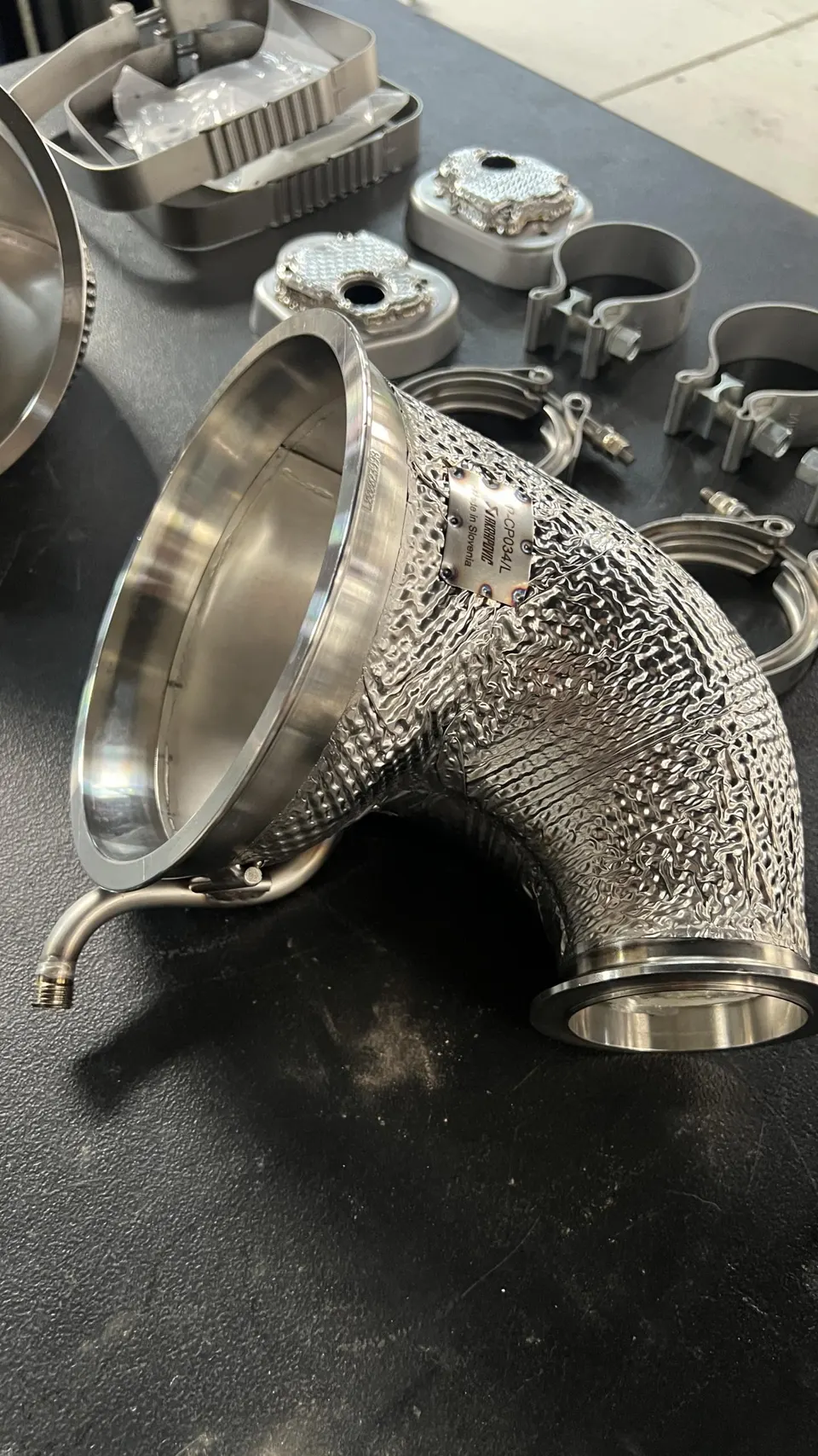 Close-up of an Akrapovic exhaust downpipe with heat shielding and other metal components on a workbench.