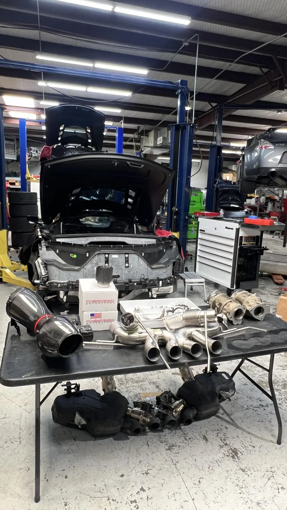 Performance exhaust and carbon fiber intake parts laid out in a shop with a car undergoing a build on a lift.