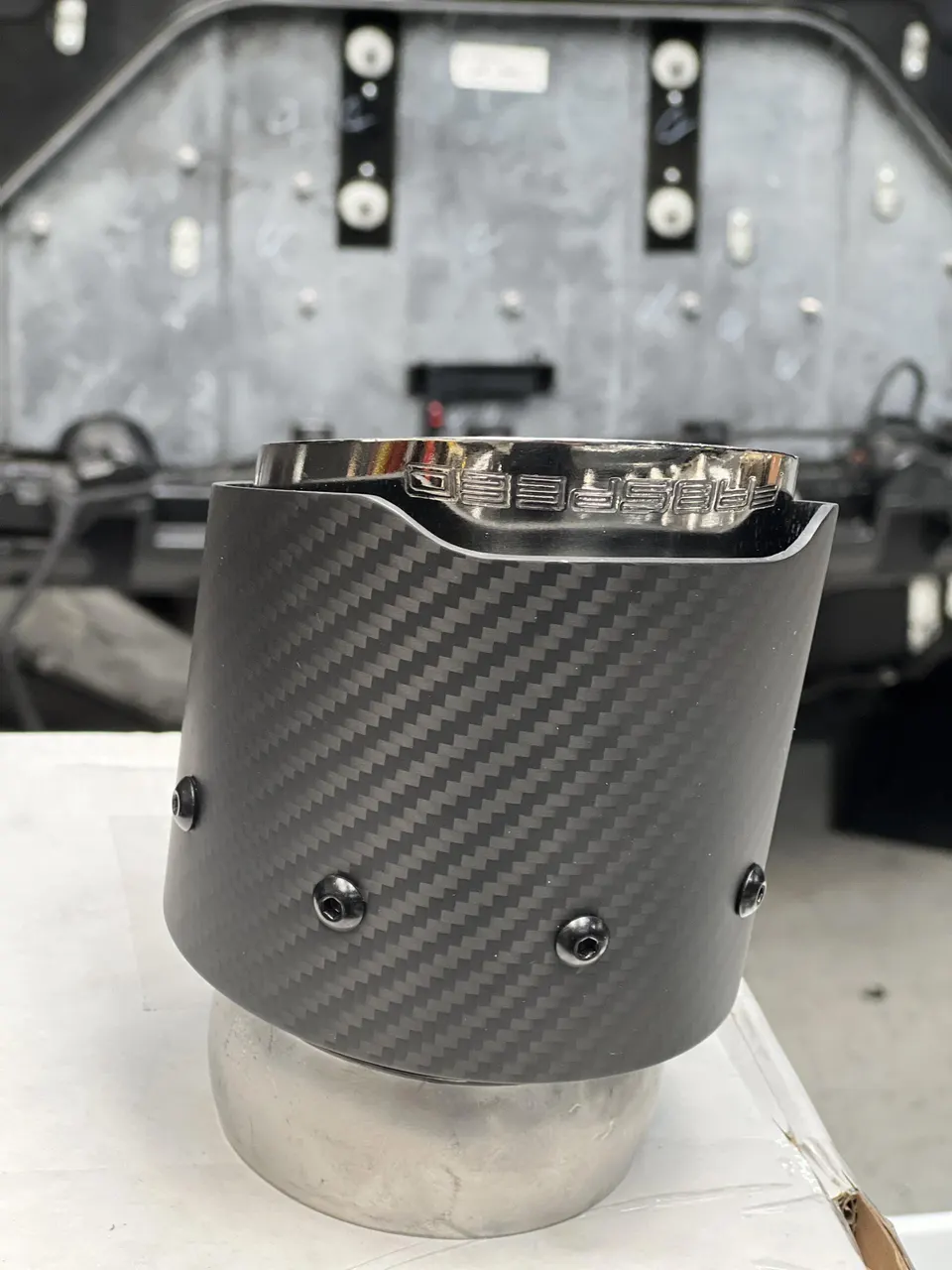 Close-up of a carbon fiber exhaust tip with a polished metal inner sleeve, likely for a high-performance vehicle, in a workshop.