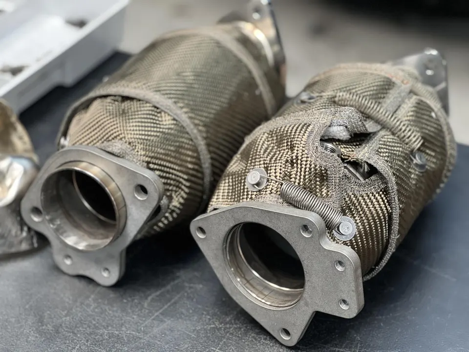 Close-up of two performance exhaust downpipes with heat wraps on a workbench, ready for installation.